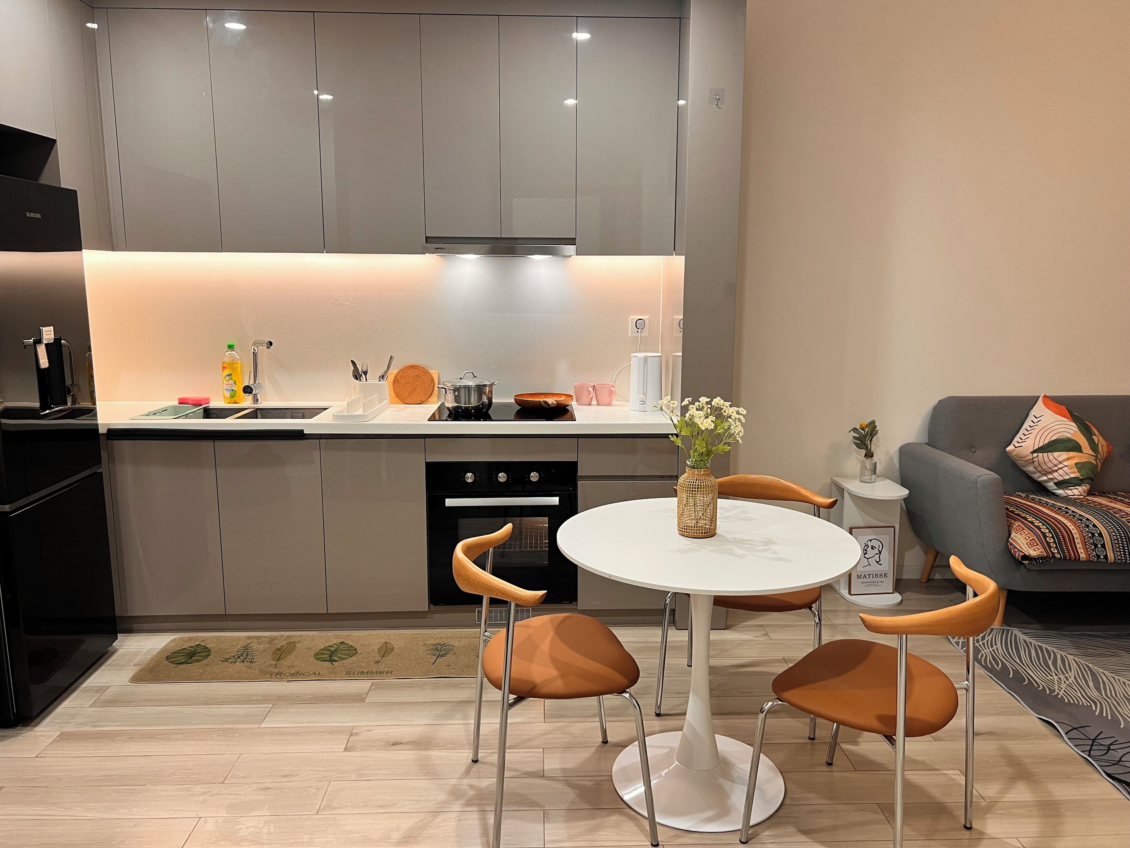 A modern kitchen with a central island, a dining area with a round table and chairs, and a grey sofa with patterned cushions.