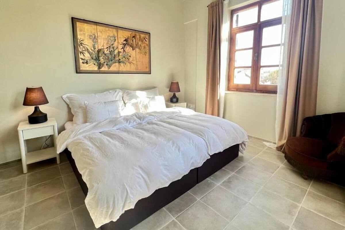 Un dormitorio con cama, dos mesitas de noche, lámpara, un cuadro, ventana con cortinas y una silla.