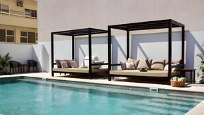 Een zwembad met twee ligbedden onder houten pergola's, een helder zwembad en een gebouw op de achtergrond.