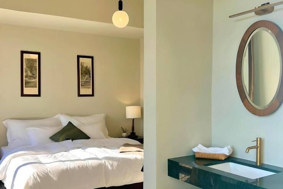 Una habitación de hotel con una cama, dos cuadros en la pared, una lámpara de noche, un espejo y un lavabo con un perchero para toallas.