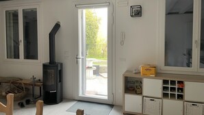 Een kamer met een houtkachel, een schuifpui naar buiten, een witte kast met wijnflessen en een houten bank.