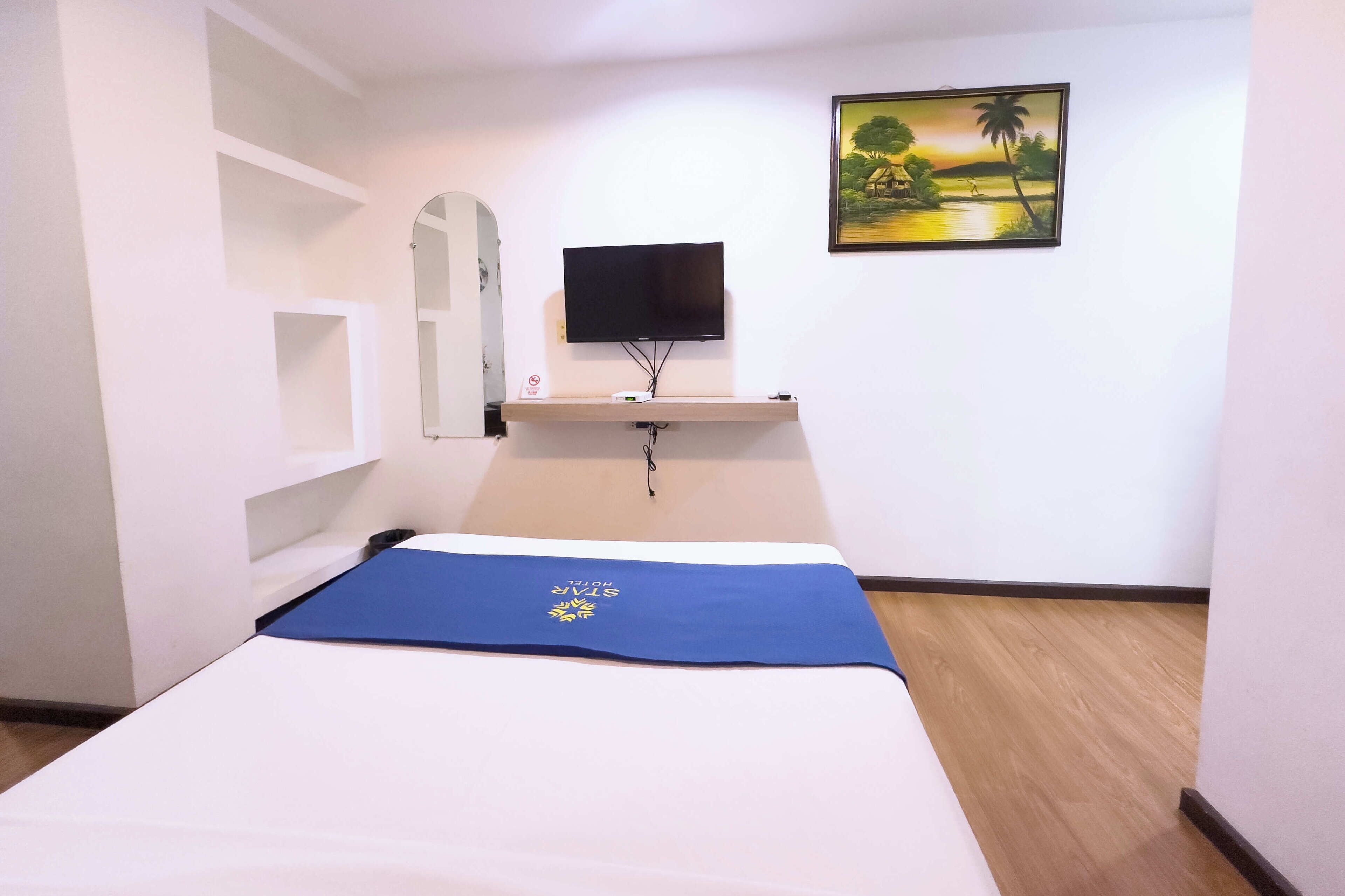 A hotel room with a bed, a TV mounted on the wall, a painting, and a blue mat on the bed.
