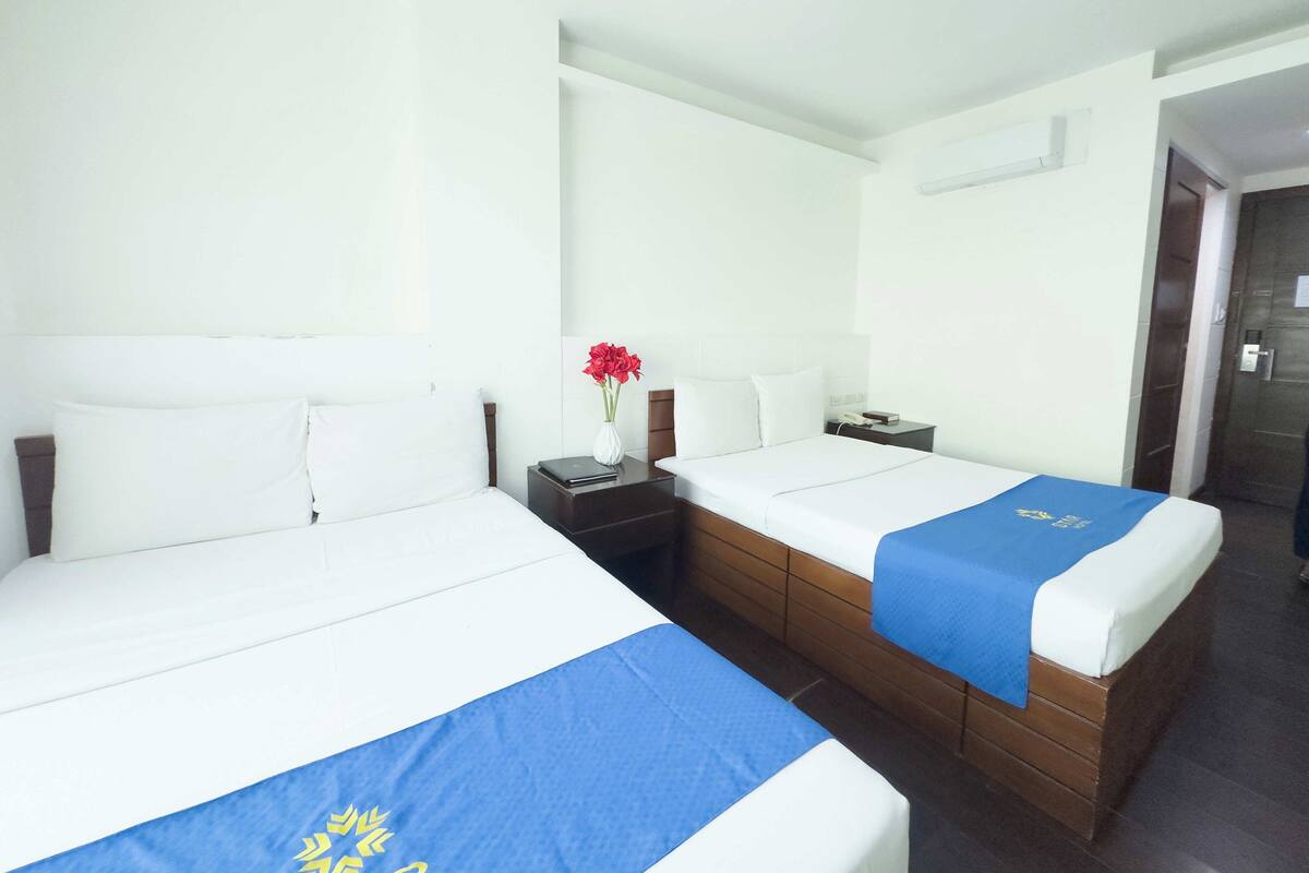 A hotel room with two single beds, a nightstand with a lamp, a vase with flowers, and a door leading to another room.