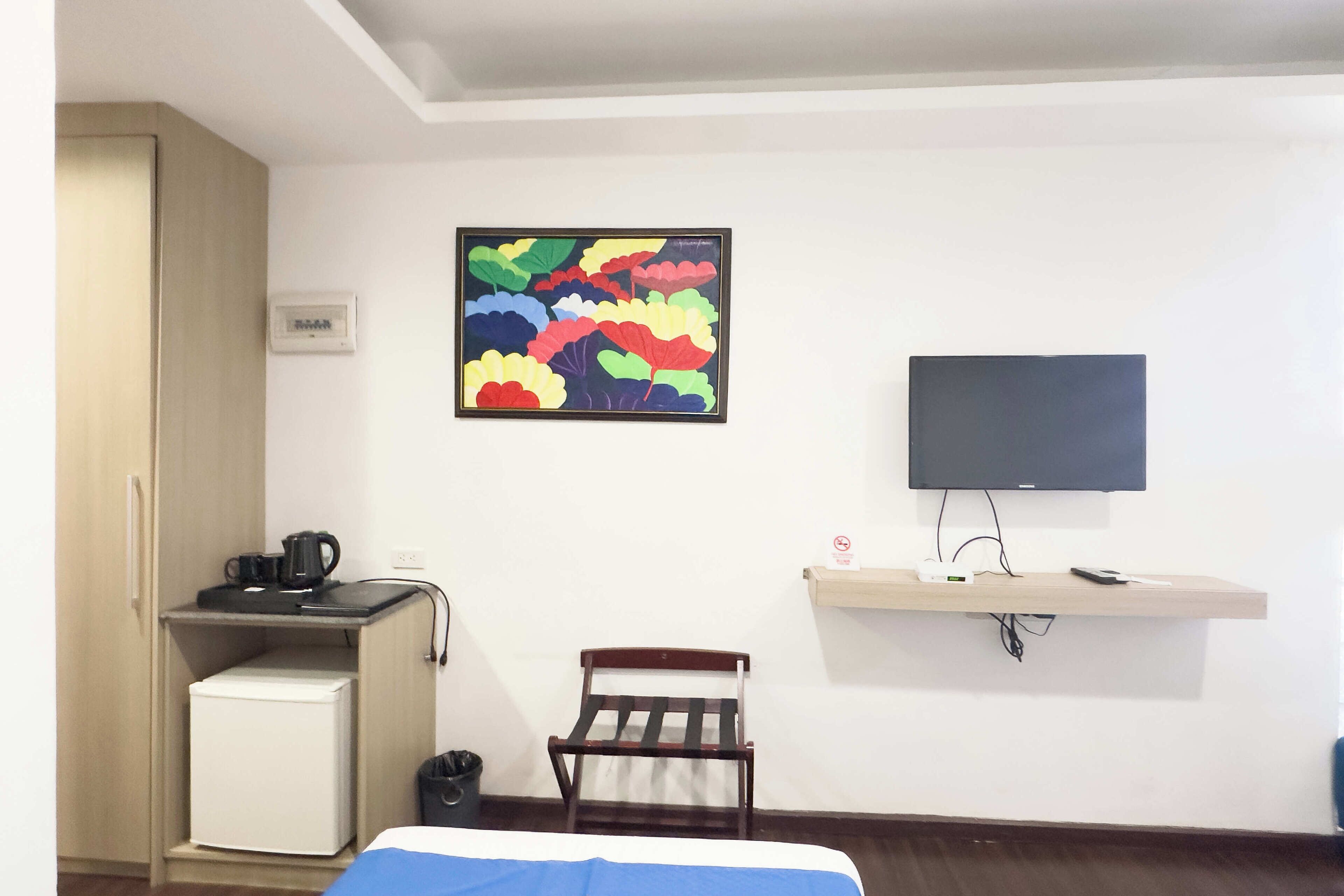 A hotel room with a bed, a desk with a kettle, a small fridge, a chair, a TV on a stand, and a colorful artwork on the wall.