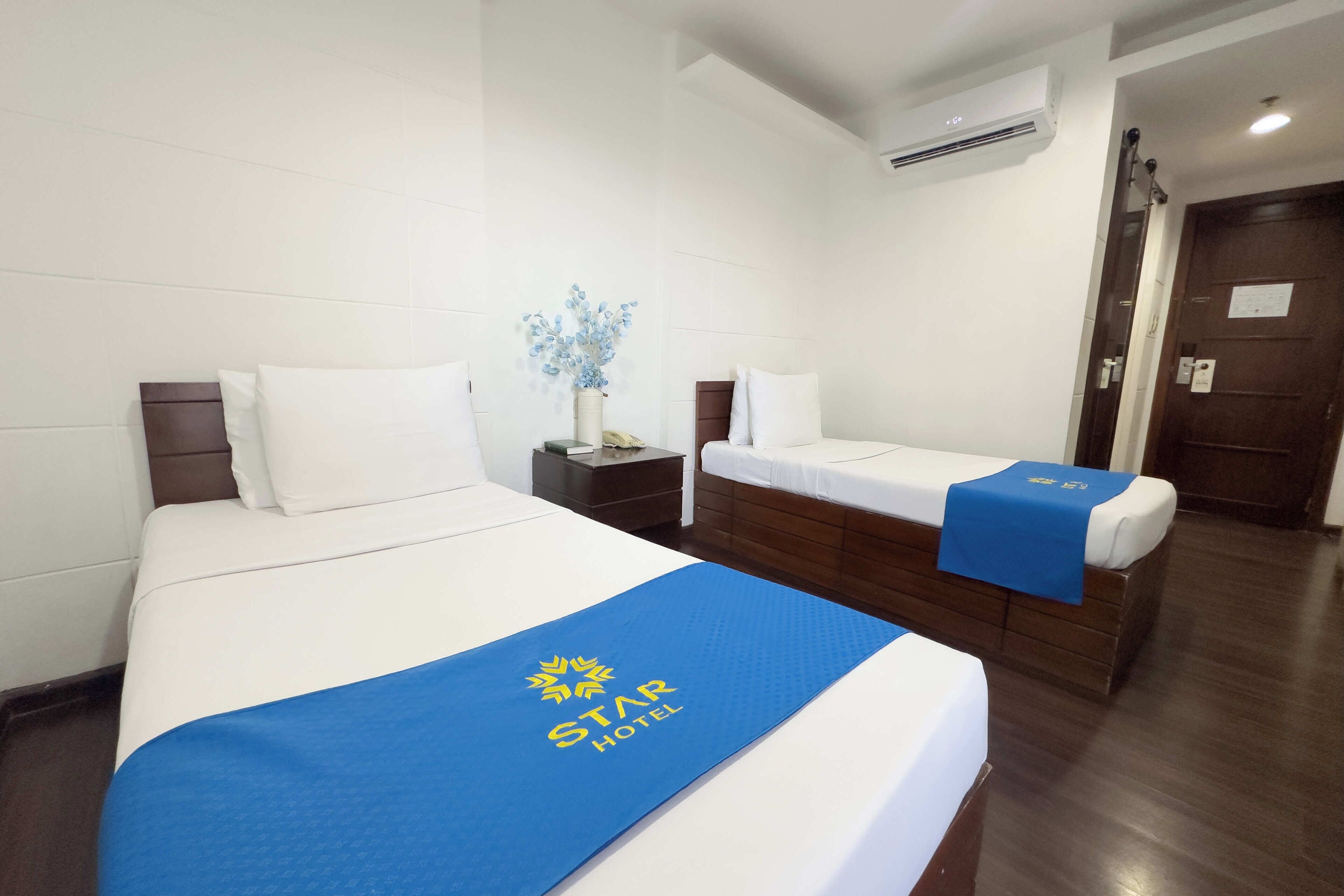 Ein Hotelzimmer mit zwei Betten, einem Nachttisch, einer Vase mit Blumen und einer blauen Bettdecke mit dem Hotel-Logo.