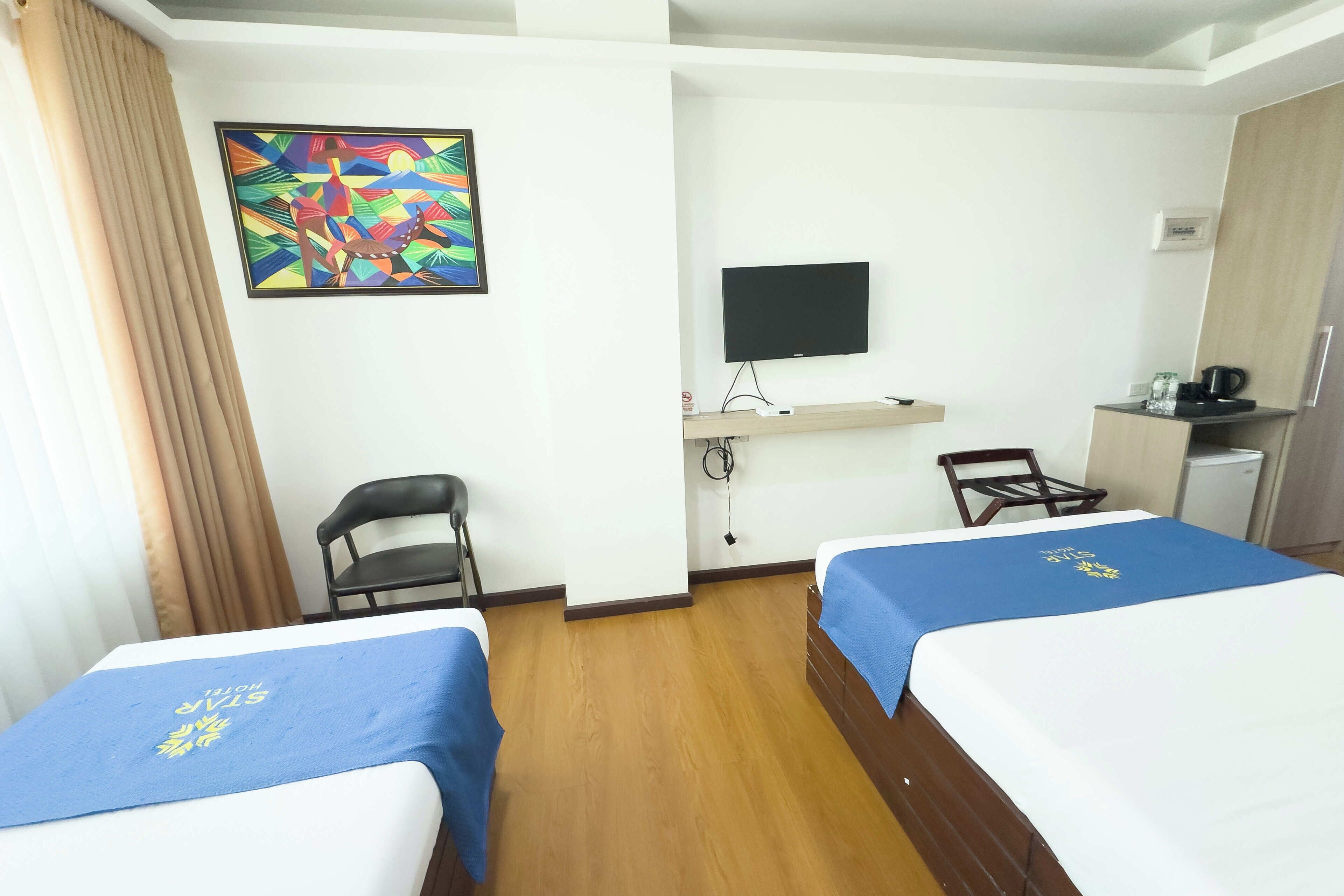 A hotel room with two beds, a TV, a desk with a computer, a chair, and a small kitchenette.
