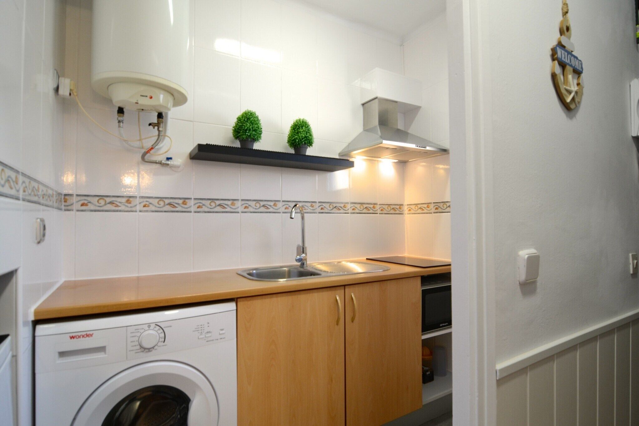Een kleine keuken met een wasmachine, houten kastjes, een roestvrijstalen spoelbak en een decoratieve plank met kleine planten.