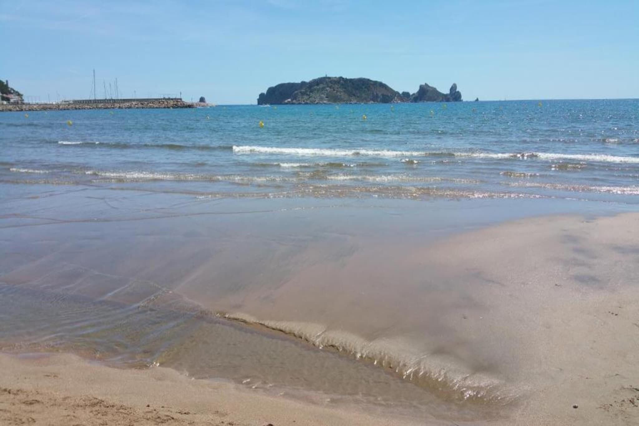 Une plage à l’eau claire, un promontoire rocheux à l’horizon et une jetée qui s’avance dans la mer.