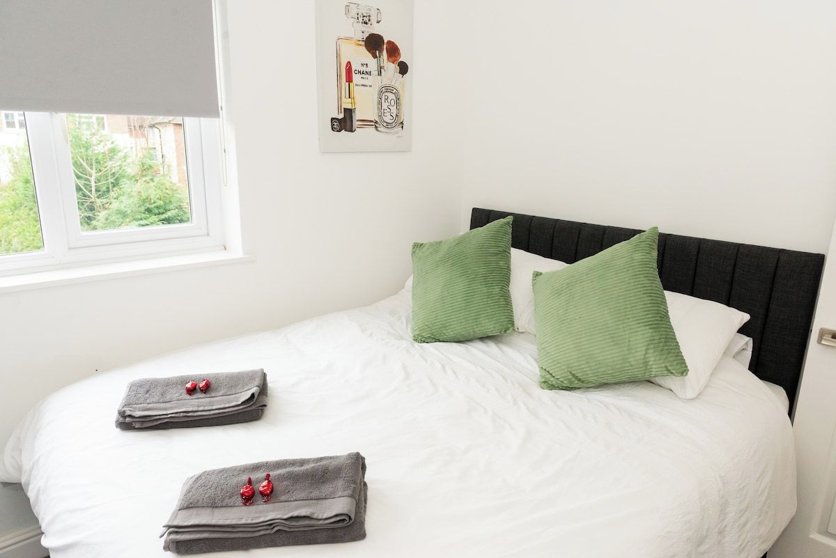 A neatly made bed with white bedding, two green pillows, and two folded towels with red accents.