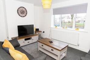 A modern living room with a flat-screen TV, a wooden coffee table, a sofa with cushions, a wall clock, and a window with a view of the outside.