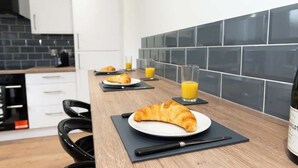 Une cuisine moderne avec un plan de travail en bois, des carreaux noirs sur le mur derrière l’évier, et un coin repas où l’on peut voir des assiettes de croissants et des verres de jus d’orange.