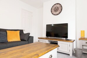 Un salon comprenant un téléviseur à écran plat, une table basse en bois, un canapé avec des coussins, une horloge murale et une petite table d’appoint avec une lampe.