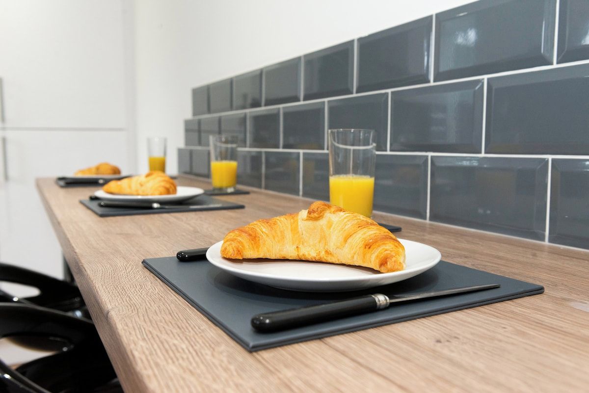 Une table en bois dressée pour le petit-déjeuner, avec des croissants sur des assiettes, un verre de jus d’orange et une serviette noire.
