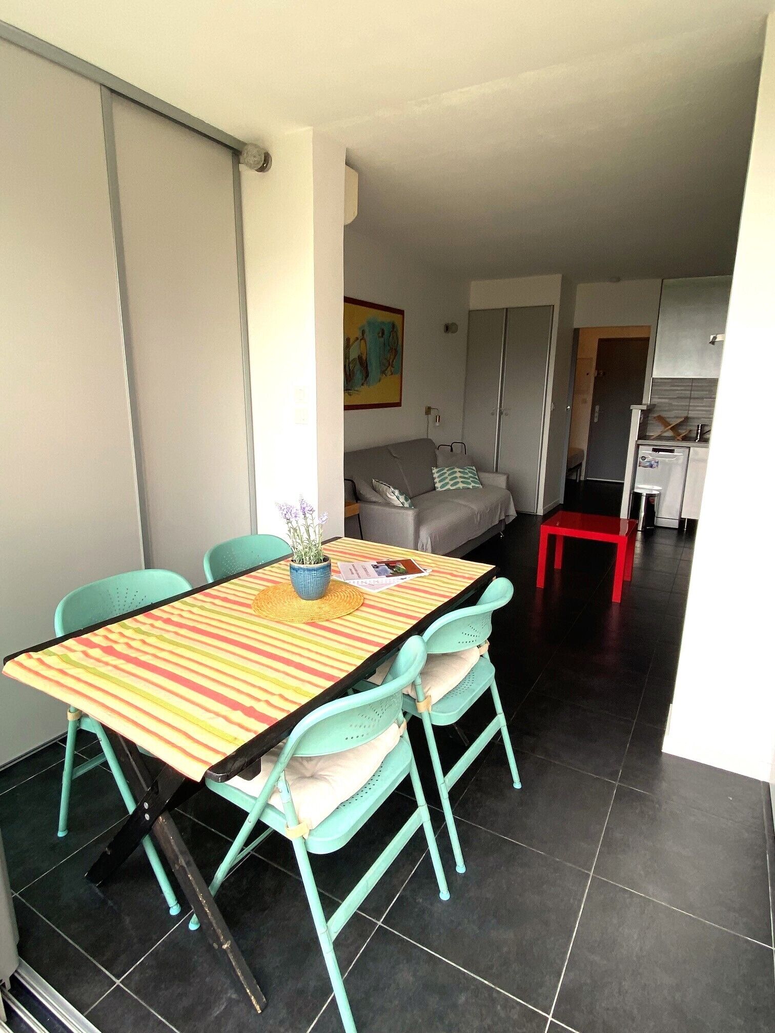 A compact living space with a dining area featuring a striped table and teal chairs, a kitchenette, and a gray sofa in the background.