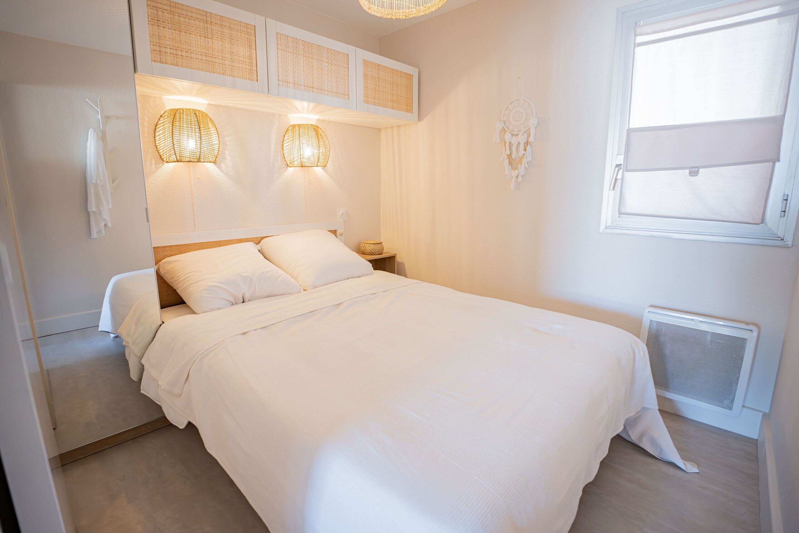 Een netjes opgemaakt bed met witte lakens, een bedlampje en een dromenvanger aan de muur.