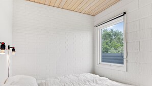 Une chambre avec un mur en briques blanches, un plafond en bois, une fenêtre donnant sur les arbres et un lit avec une literie blanche.