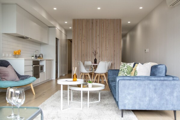 Un salon moderne avec un canapé bleu, une table basse blanche et un coin repas séparé par une cloison en bois.