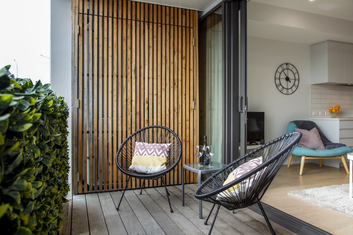 A modern living space with a wooden slatted wall, a glass table with chairs, and a kitchen area visible through an open door.