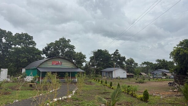 Entorno rural con un edificio de techo verde, un camino de tierra y varias construcciones más pequeñas al fondo.
