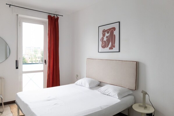 A bedroom with a bed, a window with red curtains, a framed artwork on the wall, and a small bedside table with a lamp.