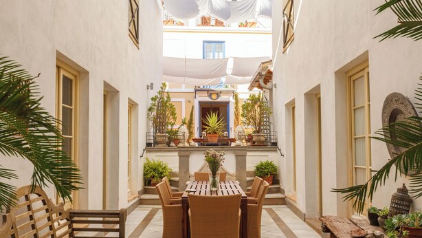 Ein Innenhof mit einer hölzernen Bank, Korbstühlen und einem Tisch mit Blumen.'