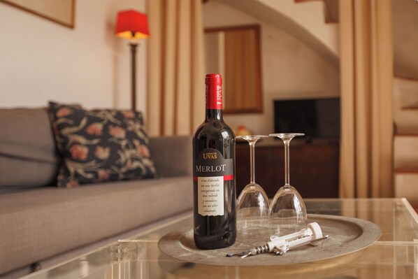 Una botella de vino Merlot, dos copas de vino y un destornillador de corcho sobre una mesa de vidrio en una sala de estar con un sofá y una lámpara al fondo.