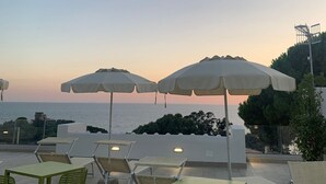 Un espacio al aire libre con mesas y sillas, grandes sombrillas y vistas al mar al atardecer.