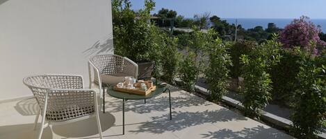 A balcony with two wicker chairs, a small table with a tray of items, and a view of the sea and greenery.