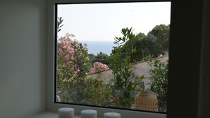 Una ventana con tres velas sobre un alféizar, que da a un jardín con flores rosadas y vistas al mar.