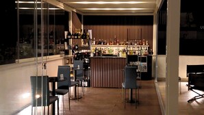 Zona de bar moderna con mostrador de madera, estanterías llenas de botellas y un espacio de estar con sillas.