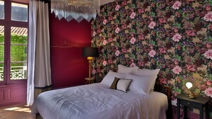 Un dormitorio con una cama grande, una araña, un papel tapiz floral y vistas a través de una ventana.