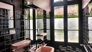 Un baño con suelo de baldosas blancas y negras en damero, un lavabo rosa y una ventana con vistas a la vegetación exterior.