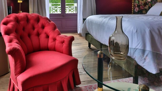 Habitación de hotel con un sillón rojo, una cama con cubrecama blanco y una mesa de centro de vidrio, con vistas a zonas verdes al exterior.