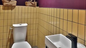 Ein Badezimmer mit Toilette, Waschbecken mit schwarzem Wasserhahn und Mülleimer. Darüber befindet sich ein Regal mit zwei gewebten Körben.