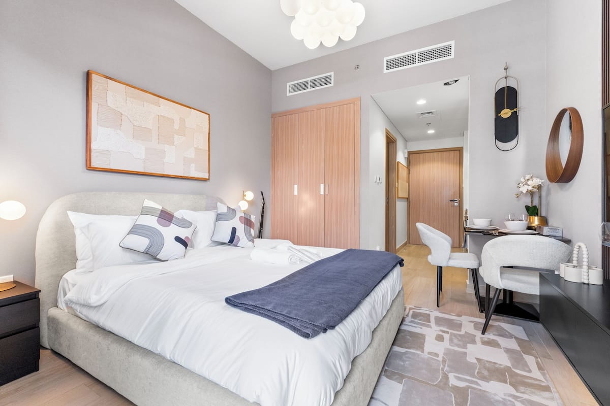 A modern bedroom with a large bed, a desk, and a chair. There is a mirror on the wall and a framed picture above the bed.