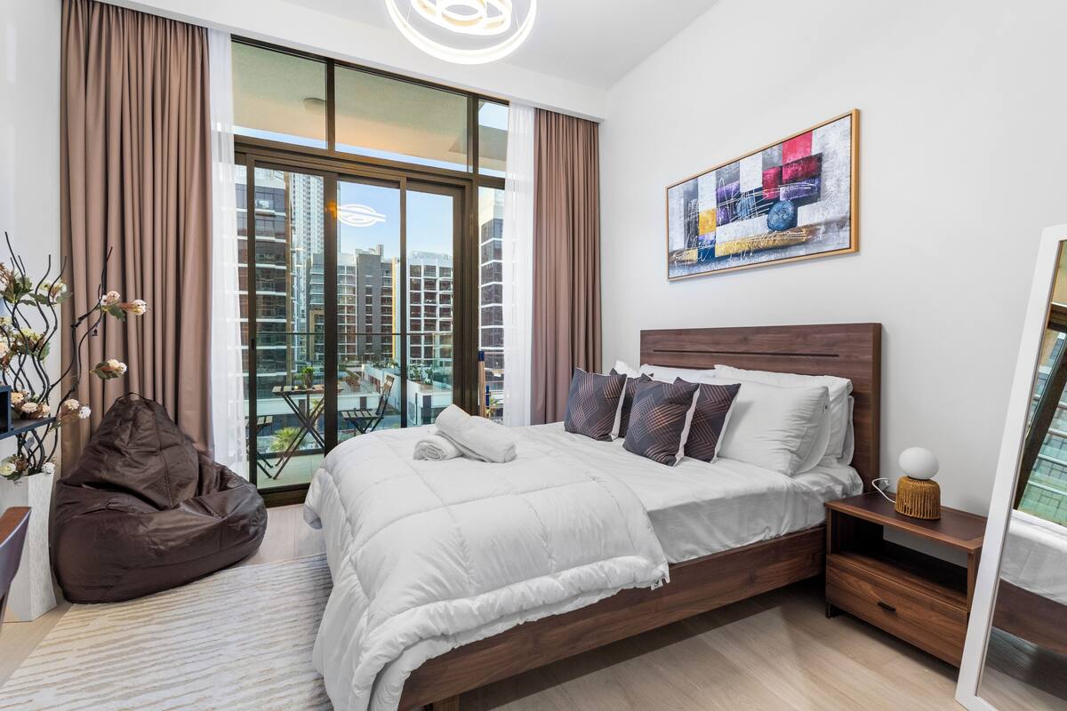 A modern bedroom with a large bed, a bean bag chair, a bedside table with a lamp, and a city view through floor-to-ceiling windows.