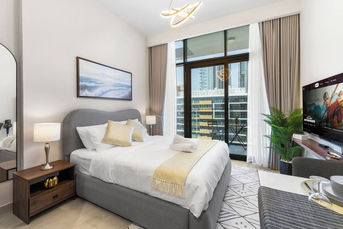 A modern hotel room with a large bed, a desk, a television, and a view of the cityscape.
