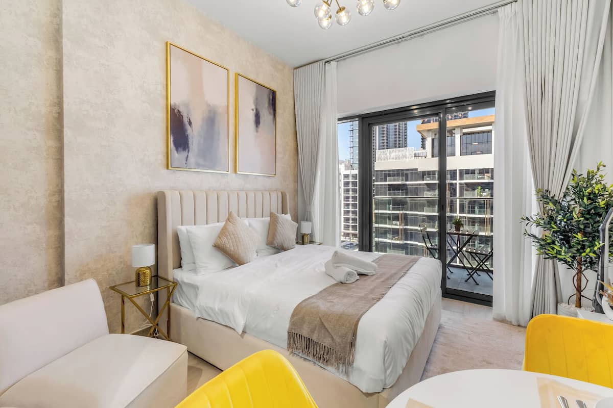 A modern hotel room with a large bed, a yellow chair, a small table, and a balcony with a view of the city.