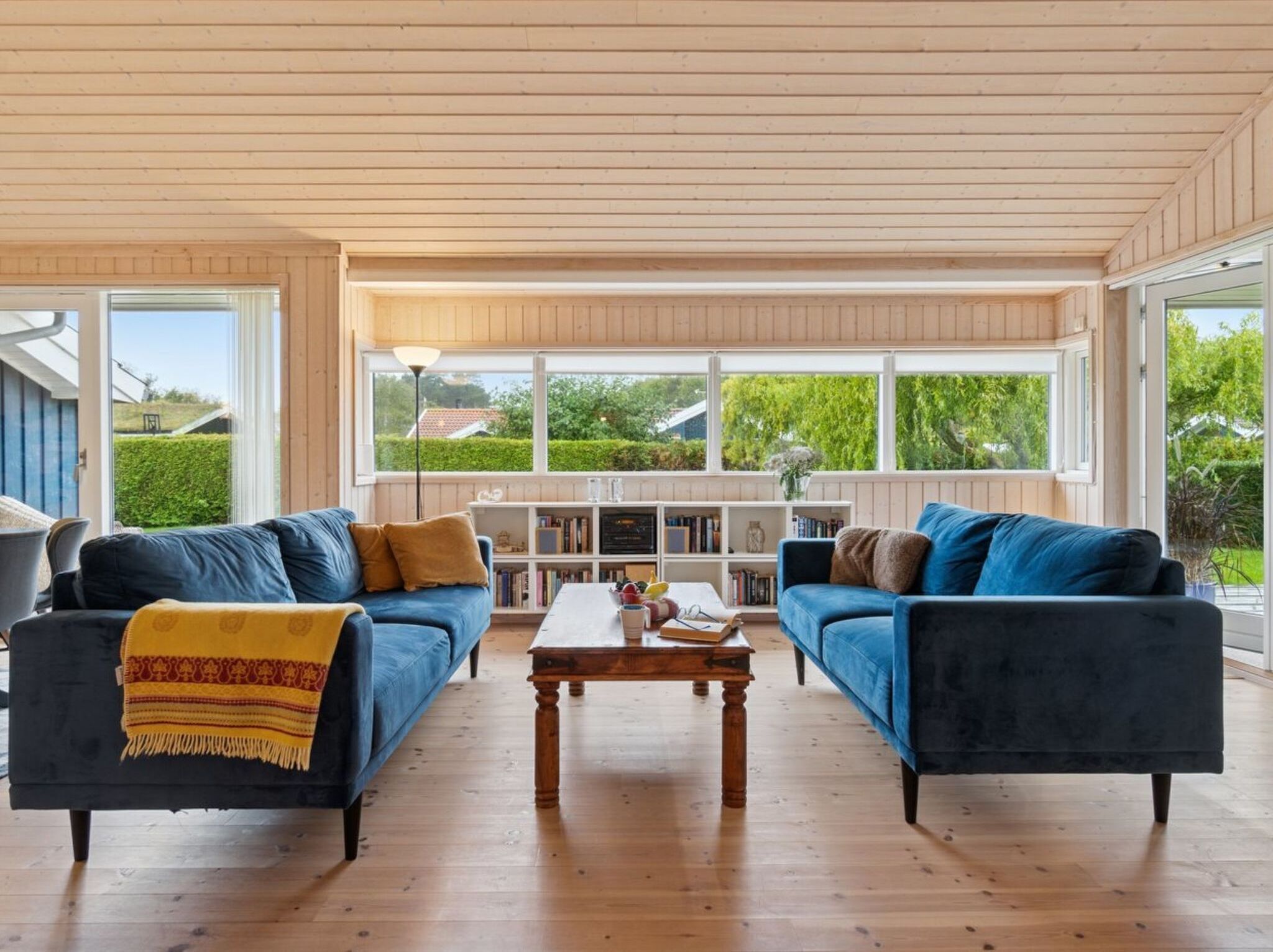 A spacious living room with two dark blue sofas, a wooden coffee table, and a bookshelf filled with books. Large windows provide a view of the outdoors.