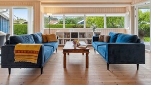 A spacious living room with two dark blue sofas, a wooden coffee table, and a bookshelf filled with books. Large windows provide a view of the outdoors.