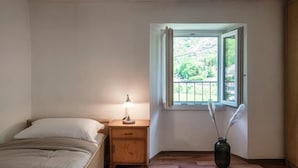 Quarto com cama, mesa de cabeceira, abajur, janela com cortinas e um vaso com penas.