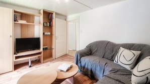 Uma sala de estar moderna com um sofá cinzento, uma televisão de ecrã plano sobre um suporte de madeira, uma pequena mesa redonda com um livro e estantes de madeira embutidas com livros e objetos decorativos.