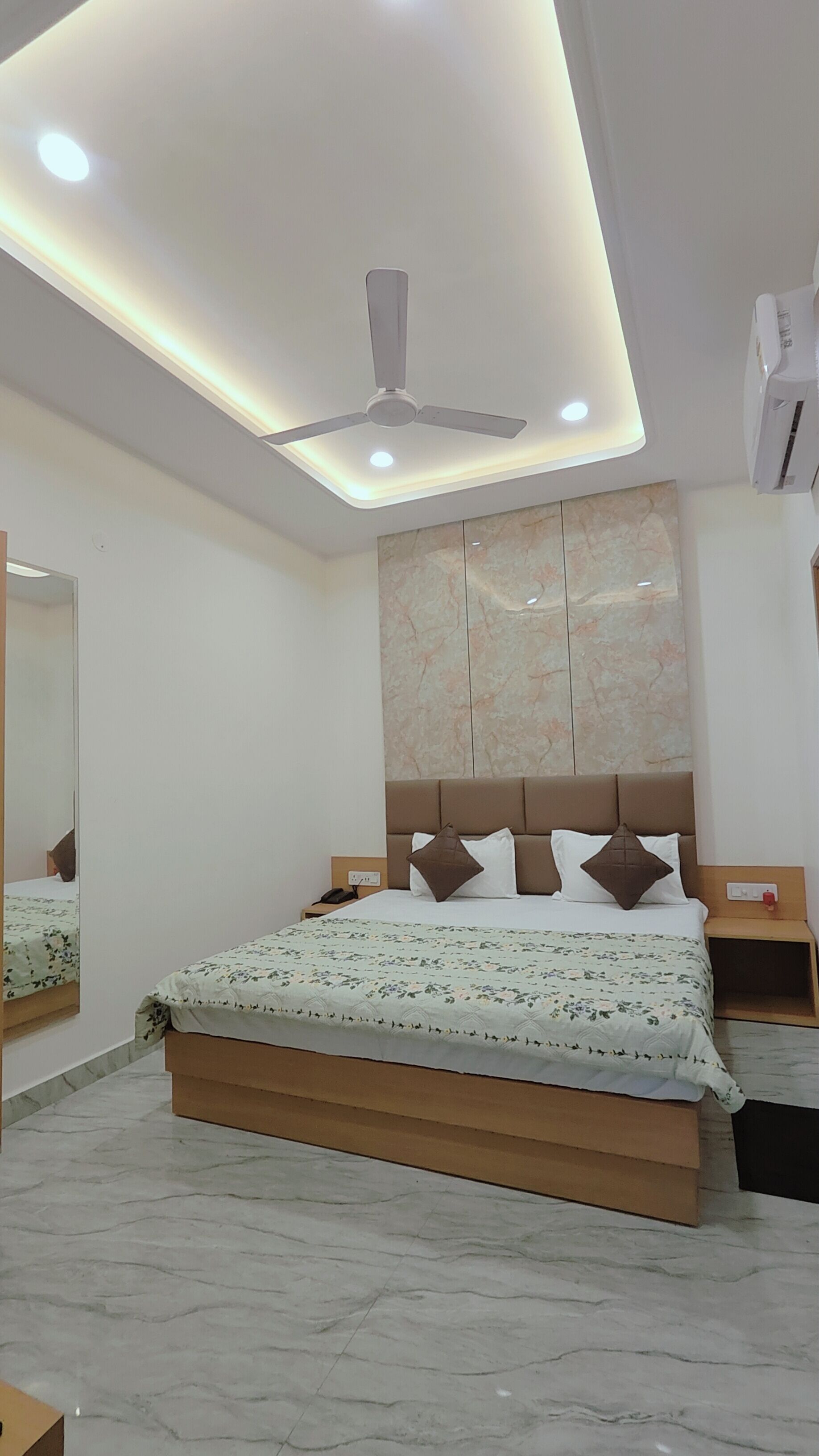 Un dormitorio moderno con cama, ventilador de techo, iluminación empotrada, piso de mármol y un espejo grande.