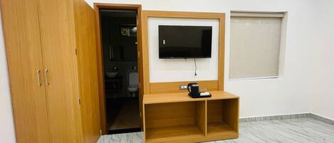 Une pièce avec une armoire en bois, un téléviseur fixé au mur, une machine à café posée sur une étagère en bois et une fenêtre avec des stores.