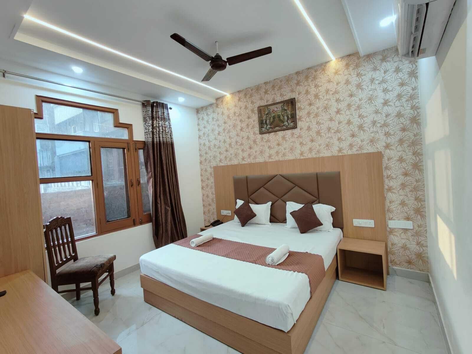 A hotel room with a large bed, a ceiling fan, a window with curtains, a wooden desk, and a small chair.