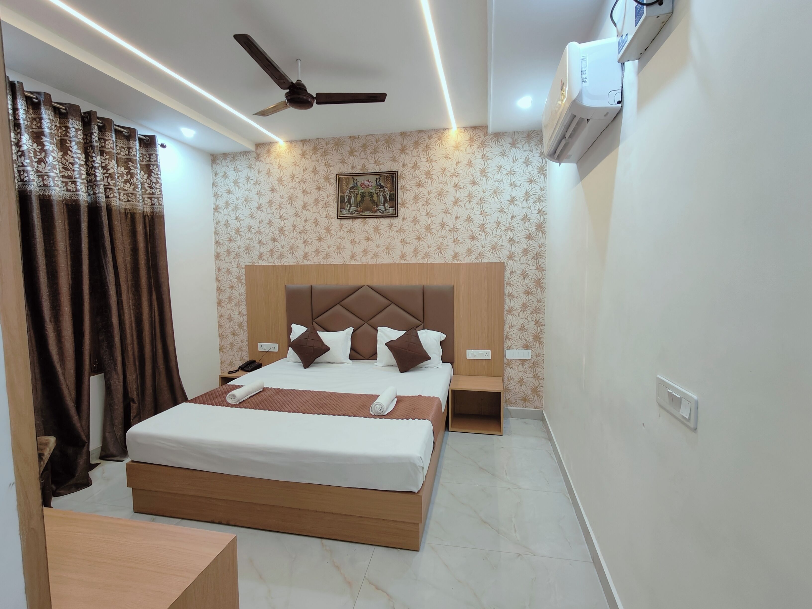 A hotel room with a bed, bedside tables, a ceiling fan, an air conditioning unit, and patterned wallpaper.