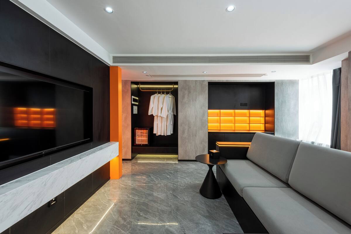 Habitación de hotel moderna con un televisor de pantalla plana grande, un sofá, una zona de estar con una mesa y un vestidor con batas colgadas.