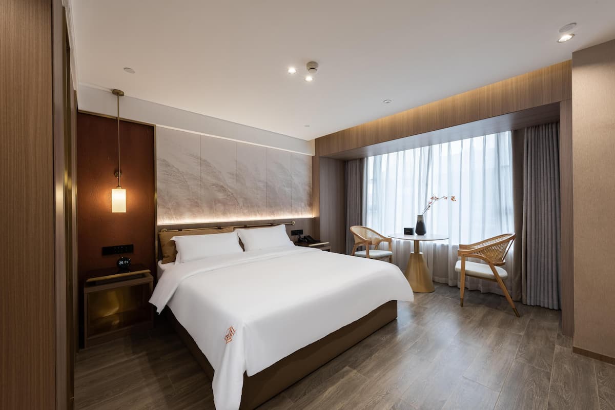 Una habitación de hotel moderna con una cama grande, mesitas de noche, una pequeña mesa redonda con sillas, un ventanal con cortinas y una lámpara de pared.