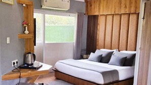 Una habitación de hotel compacta con cama, una kitchenette pequeña, una ventana con cortinas y una mesa con flores y fruta.