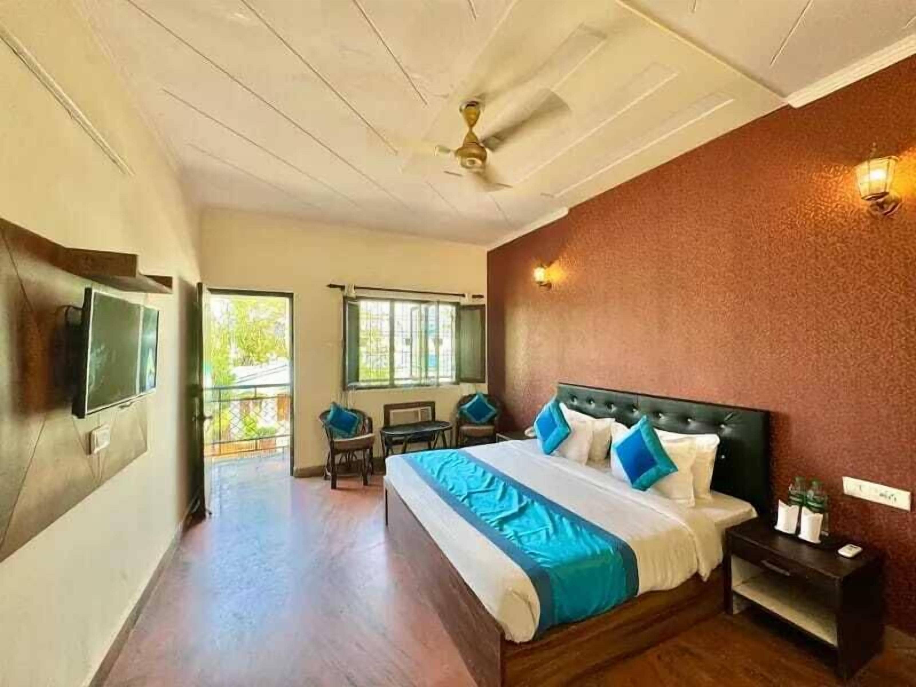 Ein Schlafzimmer mit Bett, Deckenventilator, an der Wand befestigtem Fernseher, kleinem Tisch mit Stühlen und einem Fenster mit Blick auf Grünflächen.